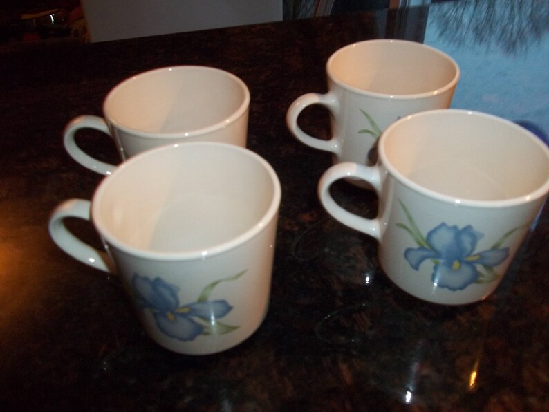 May include: Four white ceramic mugs with blue and green floral designs. The mugs have a simple, rounded shape and are perfect for everyday use.