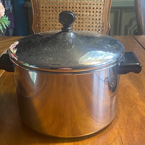 May include: A stainless steel cooking pot with a black lid and handles. The pot is sitting on a wooden table. The lid has a black knob on top. The pot is round and has a shiny surface.