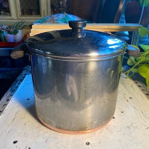 May include: A stainless steel stockpot with a copper-colored base and a matching lid. The pot has two dark-colored handles and a black knob on the lid. The pot is sitting on a white surface.