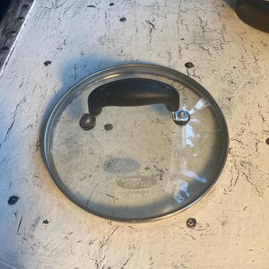 May include: A clear glass lid with a black handle and a silver rim. The lid has the words "Kitchen Essentials" and "Cuisinart" printed on it.