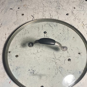 May include: A clear glass pot lid with a stainless steel rim and a black handle. The lid is circular and sits on a distressed white surface with dark spots.