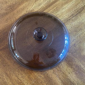 May include: A transparent brown glass lid with a small, round knob in the center. The lid is circular and appears to be for a pot or pan. The glass has a slight tint and reflects the surrounding light.