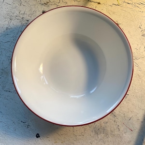 May include: A white enamel bowl with a red rim. The bowl is empty and sitting on a wooden surface.