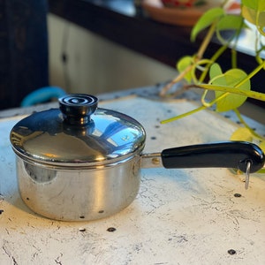 May include: A silver saucepan with a black handle and lid. The lid has a black knob. The saucepan is sitting on a white surface.