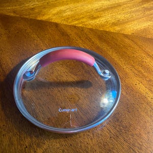May include: A clear glass Cuisinart lid with a stainless steel rim and a red handle. The lid is round and appears to be for a pot or pan. The handle is curved for easy gripping.