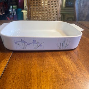 May include: Rectangular white baking dish with a lavender floral design featuring irises. The dish has a light purple border and two handles. The baking dish is on a wooden table.