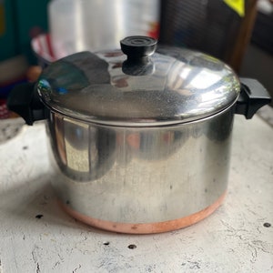 May include: Stainless steel cooking pot with a lid. The pot has a copper-colored band around the base and black handles. The lid has a black knob. The pot is sitting on a white surface.