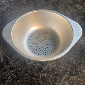 May include: A silver colander with a handle on each side. The colander has a round bottom and a mesh strainer.