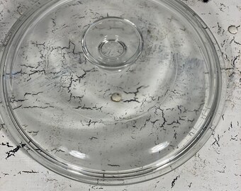 May include: A circular, clear glass lid featuring a raised, centered knob handle. The lid has a smooth, transparent surface with a defined outer rim designed to fit securely onto a corresponding container or cookware piece.