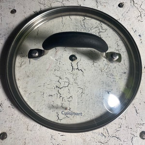 May include: A clear glass lid with a silver rim and a black handle. The lid has the word "Cuisinart" printed on it.