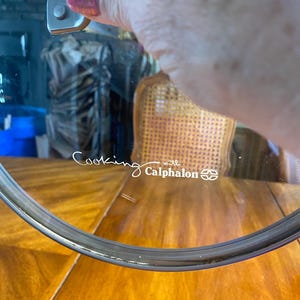 May include: A clear glass lid with a silver rim, featuring the text "Cooking with Calphalon" in a cursive font. The lid is held by a silver handle. The background includes a wooden table and a wicker chair.