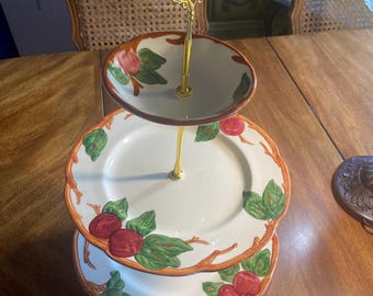 May include: A three-tiered serving stand featuring white ceramic plates with scalloped edges, decorated with painted red apples and green leaves on brown branches. The central support rod is gold-toned metal with a decorative loop handle.