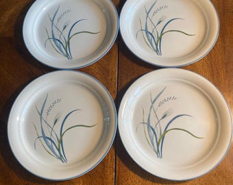 May include: A set of four round, off-white ceramic plates, each featuring a delicate, stylized botanical illustration of wheat stalks in shades of blue and green. Each plate has a simple, thin blue rim around the edge, creating a uniform look.