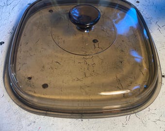 May include: A clear glass lid with a brown knob. The lid is oval shaped and has a slightly raised edge.