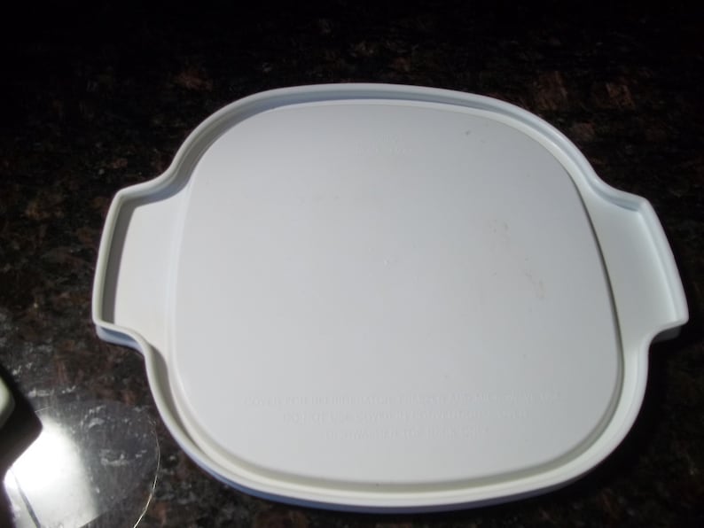 May include: A white, square-shaped plastic lid with rounded corners and two handles. The lid has a slightly raised edge and is designed to fit a container. The surface is smooth and clean, suggesting it's new or well-maintained.