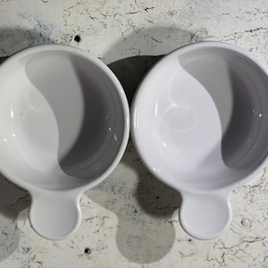 May include: Two white ceramic bowls with a handle on each side. The bowls are empty and have a smooth, glossy finish.