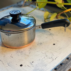 May include: A small, stainless steel saucepan with a black handle and lid. The pot has a copper-colored bottom and a black knob on the lid. The handle has a loop for hanging. The pot is sitting on a white surface.