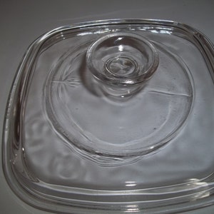 May include: A clear, square glass lid with a circular raised handle. The lid has rounded corners and a slightly textured surface. The handle is a small, circular knob. This kitchenware item is designed for use with cookware.