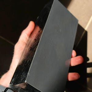 Può includere: Una pietra per affilare rettangolare nera con una superficie grigia è tenuta in mano. La pietra ha una finitura lucida e riflettente su un lato e una finitura opaca sull'altro. Viene utilizzata per affilare coltelli e utensili.