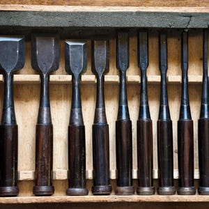 May include: A set of ten Japanese wood chisels with dark wooden handles and steel blades, arranged in a rectangular wooden storage box. The chisels vary in blade width, featuring a tapered metal neck connecting the handle to the cutting edge.