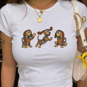 Op de afbeelding: Wit T-shirt met een geborduurd ontwerp van drie cartoon teckels. De honden zijn bruin met bruine buiken, blauwe kragen en gestreepte details. Het shirt heeft een ronde hals en korte mouwen.