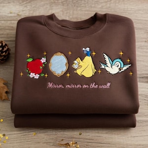May include: A folded brown sweatshirt with embroidered images of a red apple, a mirror, a woman in a yellow dress, and a blue bird. The text "Mirror, mirror on the wall" is embroidered below the images.