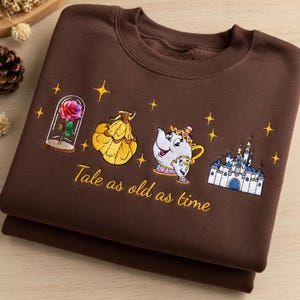 May include: A folded brown sweatshirt with embroidered images from Beauty and the Beast. The design includes a rose in a glass case, Belle's ball gown, Mrs. Potts and Chip, a castle, and the text "Tale as old as time."
