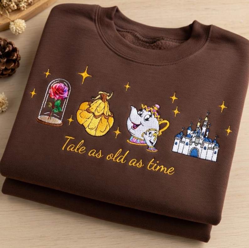 May include: A folded brown sweatshirt with embroidered images from Beauty and the Beast. The design includes a rose in a glass case, Belle's ball gown, Mrs. Potts and Chip, a castle, and the text "Tale as old as time."