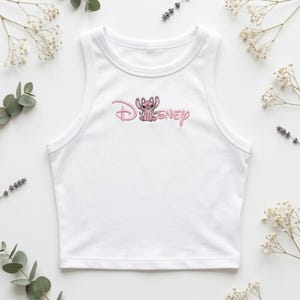 May include: White tank top with a pink embroidered Disney Stitch design. The design features the word "Disney" in a cursive font with a cartoon Stitch character. The top is sleeveless and has a rounded neckline.