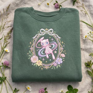 Könnte beinhalten: Ein dunkelgrünes Sweatshirt mit einem gestickten Design eines rosa Cartoon-Charakters in einem floralen und Schleifenrahmen. Das Design umfasst Pastellfarben und Sterne. Das Sweatshirt ist gefaltet und von Blumen umgeben.