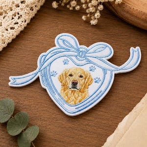 May include: A heart-shaped embroidered patch featuring a golden retriever portrait in the center. The patch is outlined in light blue thread and topped with a decorative light blue bow. Small light blue paw prints are scattered around the dog.