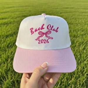 May include: A white and pink baseball cap with the text "Bach Club 2026" embroidered in pink. The cap has a curved brim and a pink button on top. The background is green grass.
