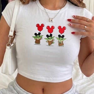 May include: White crop top featuring a cartoon graphic of three Grogu characters holding red Mickey Mouse-shaped balloons. The Grogu characters are wearing brown robes and black ears. The top has a round neckline and short sleeves.