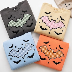 Può includere: Quattro felpe girocollo in grigio antracite, crema, azzurro e arancione. Ognuna presenta un grande pipistrello centrale con motivo a quadretti vichy verde, rosa, blu o arancione, circondato da piccoli pipistrelli e stelle nere ricamate.