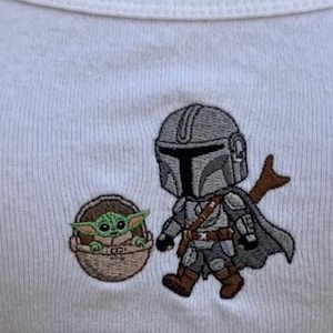 K&ouml;nnte beinhalten: Wei&szlig;es geripptes Tanktop mit gestickten Cartoon-Bildern von The Mandalorian und Grogu. Der Mandalorianer tr&auml;gt eine graue R&uuml;stung mit Umhang, und Grogu befindet sich in einer braunen Trage.