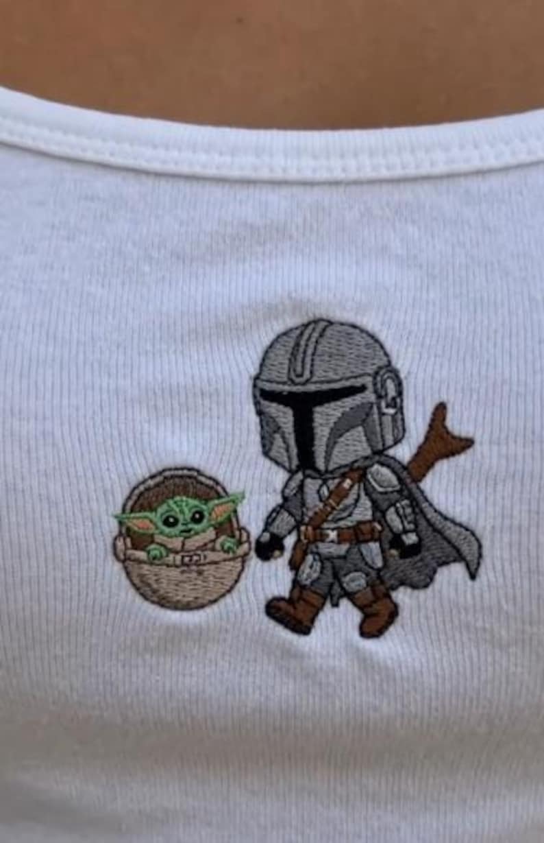 K&ouml;nnte beinhalten: Wei&szlig;es geripptes Tanktop mit gestickten Cartoon-Bildern von The Mandalorian und Grogu. Der Mandalorianer tr&auml;gt eine graue R&uuml;stung mit Umhang, und Grogu befindet sich in einer braunen Trage.