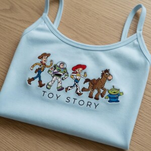 May include: Light blue tank top featuring an embroidered design of characters from the animated film "Toy Story." The design includes Woody, Buzz Lightyear, Jessie, Bullseye, and the Little Green Men. The words "TOY STORY" are embroidered below the characters.