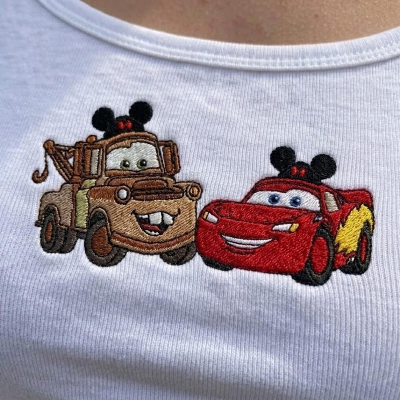 May include: A white ribbed t-shirt featuring an embroidered design of two cartoon vehicles. On the left, a brown tow truck with a hook and black mouse ears. On the right, a red race car with a yellow lightning bolt graphic and black mouse ears.