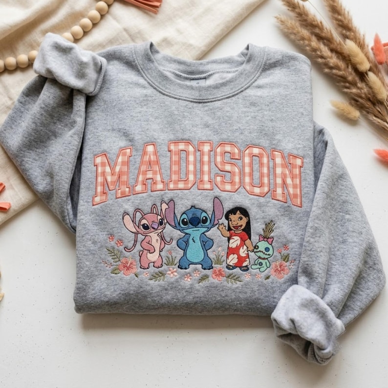 Puede incluir: Una sudadera gris jaspeado de cuello redondo con el nombre MADISON en letras grandes de bloque con estampado de cuadros vichy naranja y blanco. Debajo del texto hay un bordado de Angel, Stitch, Lilo y Scrump rodeados de peque&ntilde;as flores.