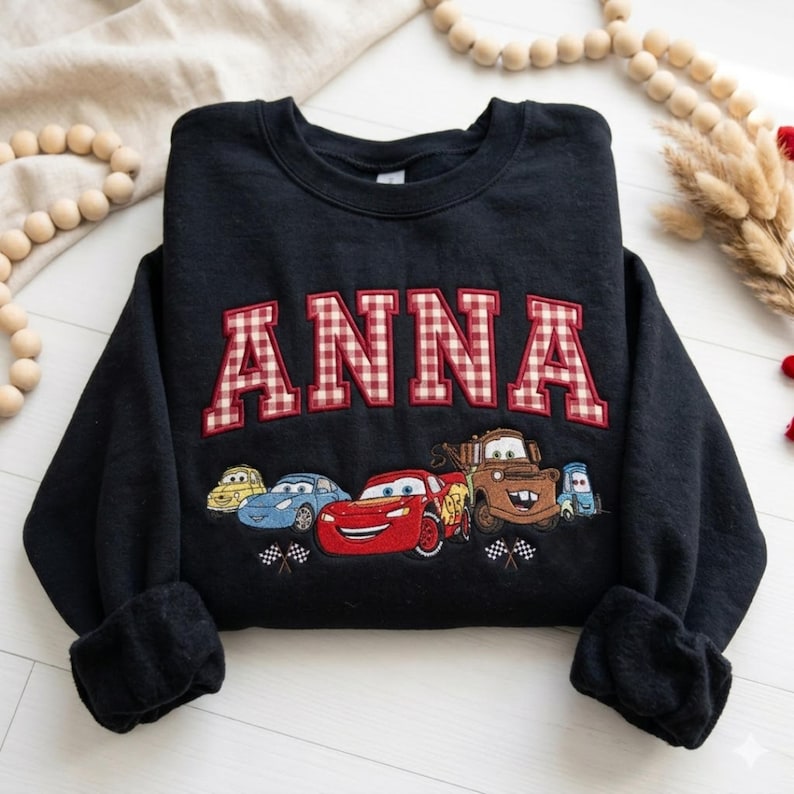 May include: A black long-sleeve sweatshirt featuring the name ANNA in red and white gingham block letters. Below the name is an embroidered graphic of five cartoon cars, including a red race car and a brown tow truck, with checkered flags.