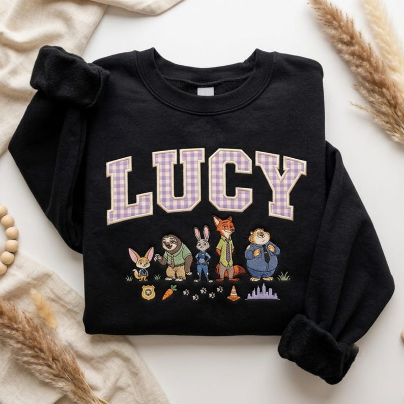 May include: A black long-sleeve sweatshirt featuring the name LUCY in large, light purple and white gingham varsity-style letters. Below the text is an embroidered graphic of five animated animal characters, a badge, a carrot, paw prints, and a cone.