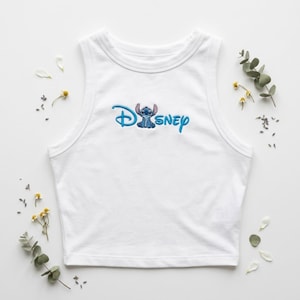 May include: White sleeveless crop top with the word "Disney" embroidered in blue, featuring the character Stitch. The top is surrounded by small white petals and sprigs of greenery.