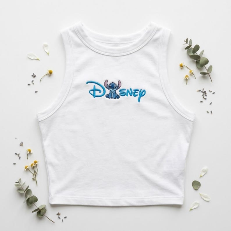 May include: White sleeveless crop top with the word "Disney" embroidered in blue, featuring the character Stitch. The top is surrounded by small white petals and sprigs of greenery.