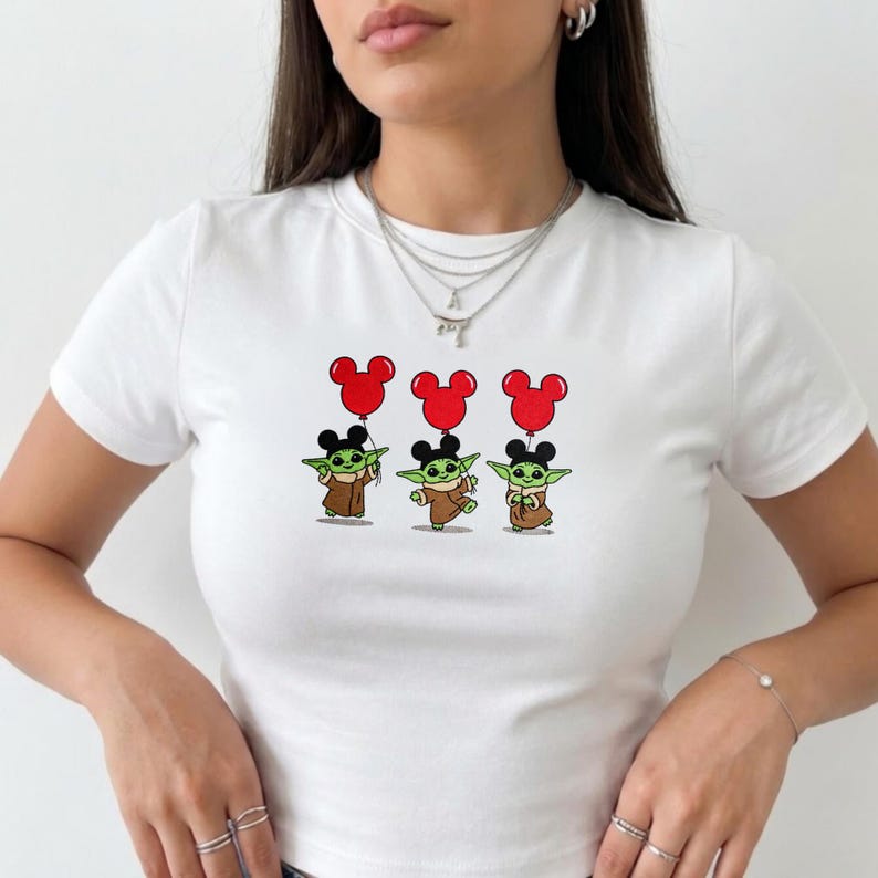 May include: White t-shirt featuring three cartoon Grogu characters holding red Mickey Mouse balloons. The Grogu characters are wearing brown robes and have black ears. The balloons are red.