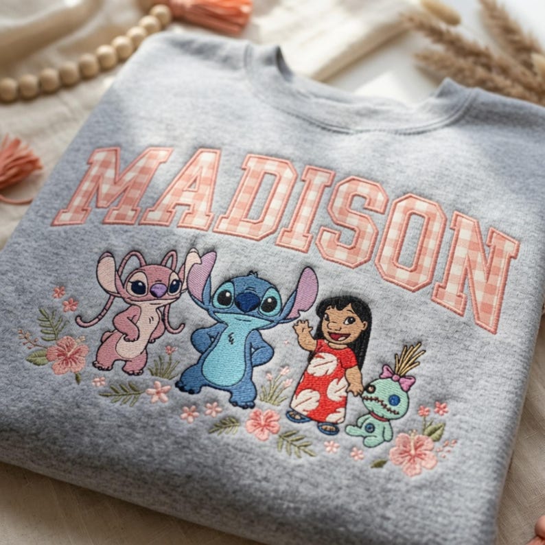 Puede incluir: Una sudadera gris jaspeado de cuello redondo con el nombre MADISON en letras de bloque a cuadros rosas y blancos. Debajo del texto hay un bordado de Angel, Stitch, Lilo y Scrump rodeados de peque&ntilde;as flores de hibisco rosas y hojas verdes.