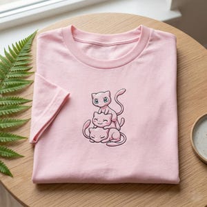 May include: A pale pink t-shirt with a cartoon illustration of three pink cat-like creatures, one on top of the other. The creatures have large eyes and long tails. The t-shirt is folded on a wooden surface.