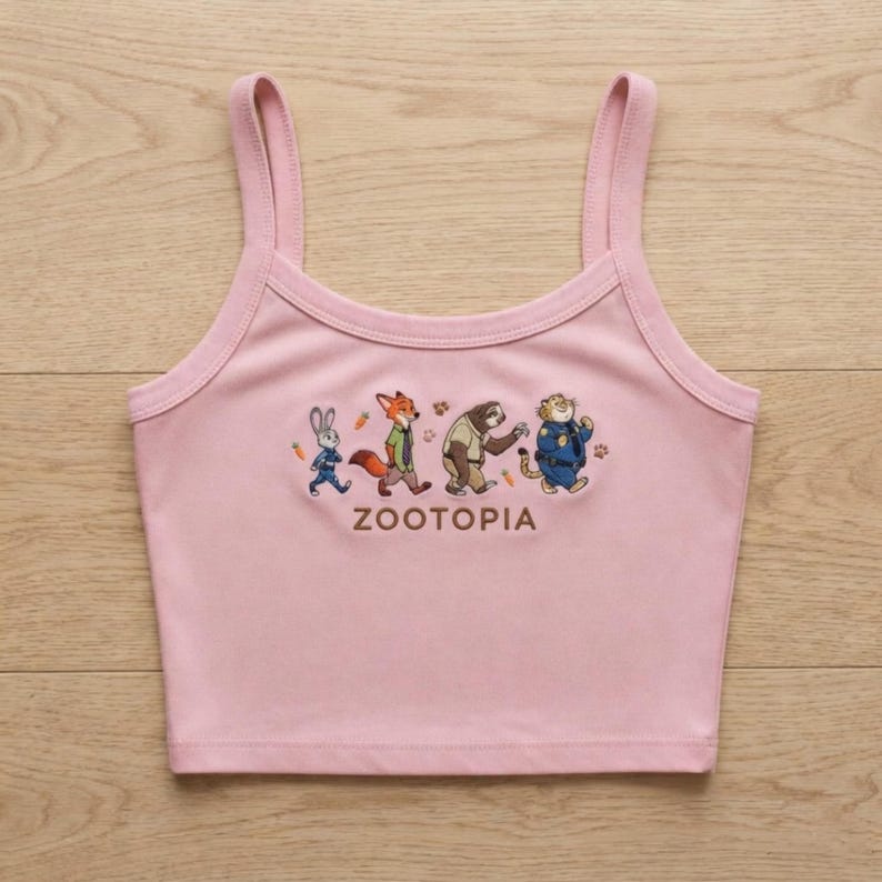 May include: A light pink tank top with thin straps. The front features a graphic of characters from the movie "Zootopia" and the word "ZOOTOPIA" in capital letters. The characters include a rabbit, fox, sloth, and tiger.