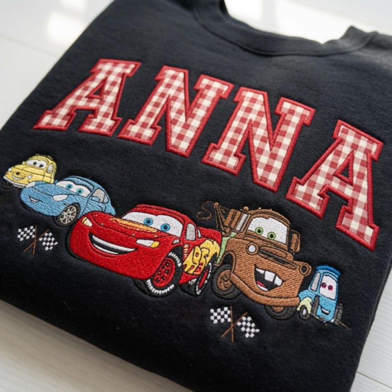 May include: A black sweatshirt featuring the name ANNA in large, red and white gingham-patterned applique letters. Below the name is an embroidered graphic of four cartoon cars, including a red race car and a brown tow truck, with checkered flags.