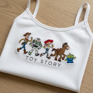 May include: White tank top with embroidered Toy Story characters: Woody, Buzz Lightyear, Jessie, Bullseye, and the Little Green Men. The text "TOY STORY" is embroidered below the characters. The top has thin straps.