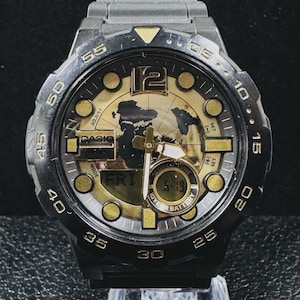 May include: A black Casio wristwatch with a gold-toned dial featuring a world map graphic. The watch has a black resin bezel with minute markers, a digital display showing 'FRI' and '6-19', analog hands, and a '10 YEAR BATTERY' label on the face.